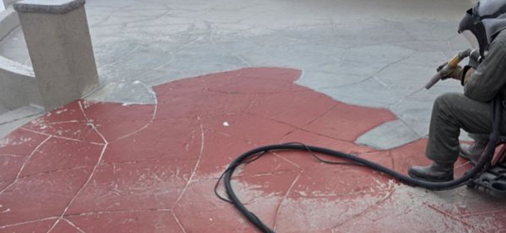 A professional technician using dustless blasting to strip red paint from a decorative stamped concrete patio, revealing the clean stone surface underneath.